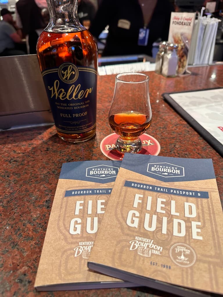Weller Full Proof bourbon, a Glencairn tasting glass, and Kentucky Bourbon Trail passports at an airport bar before a trip to Kentucky