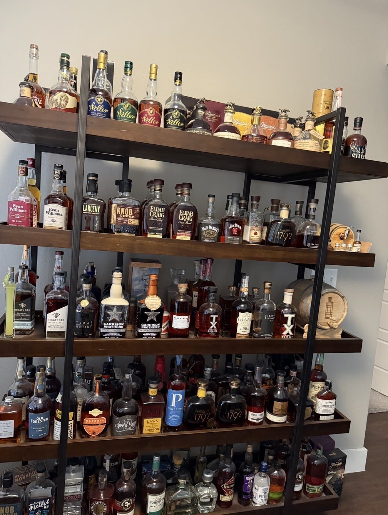 A five-shelf wood and black metal bookshelf holding more than a hundred bourbon bottles, with Weller and Blanton's bottles on the top shelf, barrel proof bottles on the second, and Texas bourbons on the third