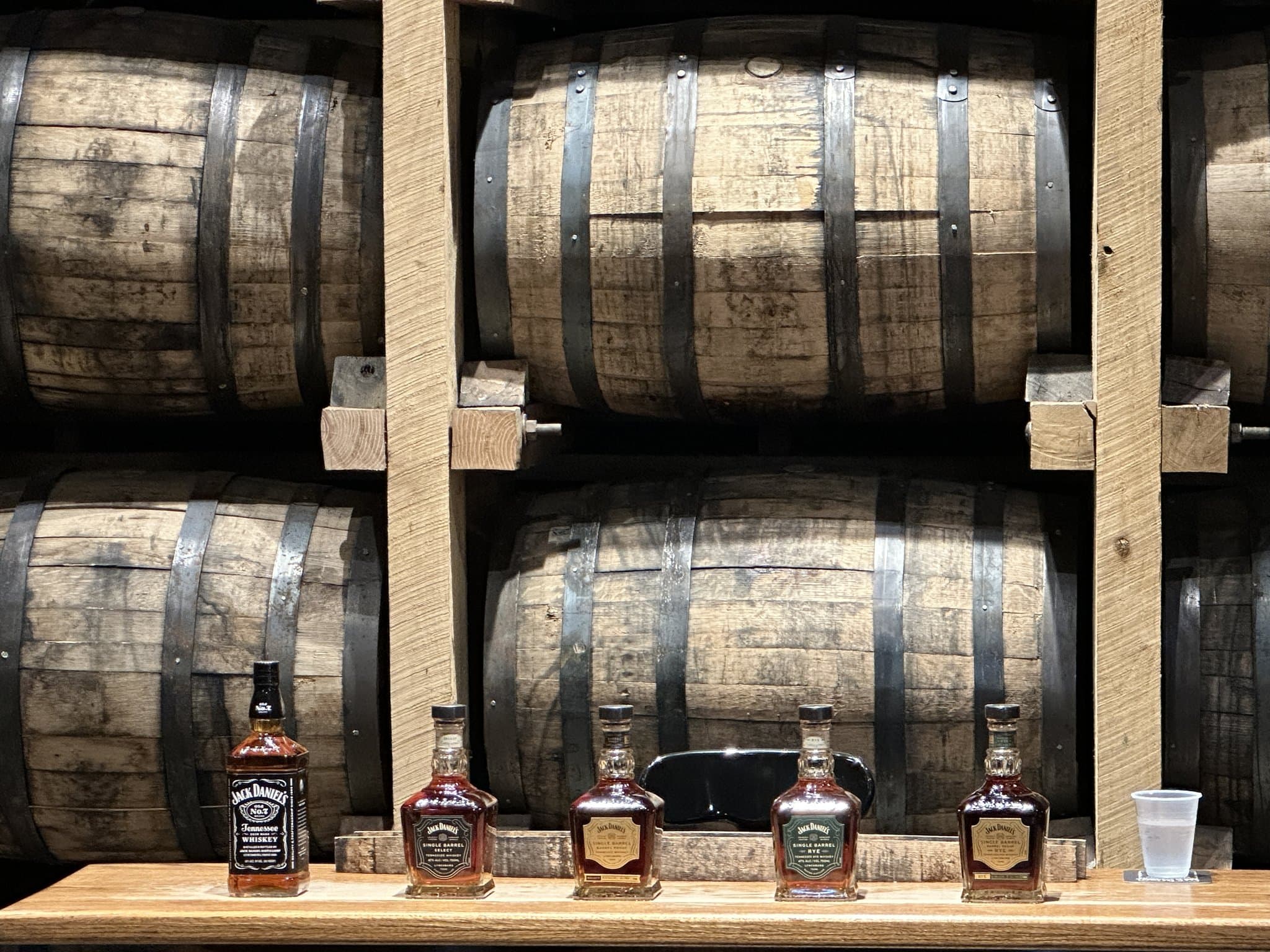 Five Jack Daniel's bottles lined up on a barrel rack in the Lynchburg rickhouse: Old No. 7, Single Barrel Select, Single Barrel Barrel Proof, Single Barrel Rye, and Single Barrel Barrel Proof Rye